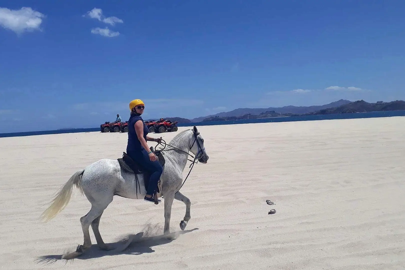 Image of a woman wearing a helmet and horseback riding on the stunning Conchal Beach. The blue ocean can be seen in the background, with clear skies and white sand creating a picturesque setting.